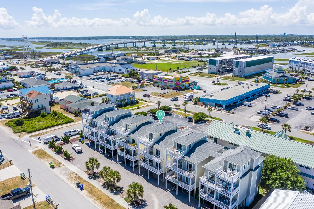 A vibrant exterior scene featuring multi-story townhouses surrounded by lush palm trees. The nearby commercial area is visible, with people relaxing on balconies, exploring local shops, and dining at restaurants. The setting sun casts a warm glow, and a bridge spans the background, hinting at walkable attractions and waterfront activities.