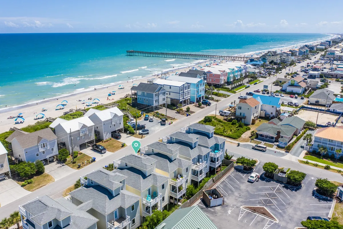 A lively beachfront scene with the iconic Surf City Pier extending into the ocean, framed by beach houses and colorful umbrellas. Swimmers and beachgoers dot the sand, and the pier hints at fishing, scenic walks, and waterfront family outings. Local shops and restaurants are visible just off the beach.