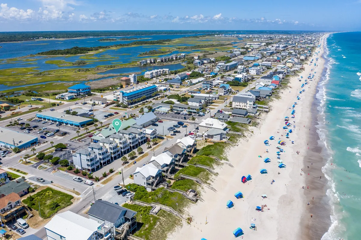 A wide exterior shot encompassing the beach, ocean, nearby houses, parked cars, and colorful umbrellas. The scene is lively yet relaxed, showing people enjoying swimming, sunbathing, and exploring local shops and seafood restaurants. The image captures sunset views over the ocean, festive beach activities, and the proximity of the neighborhood to local attractions.