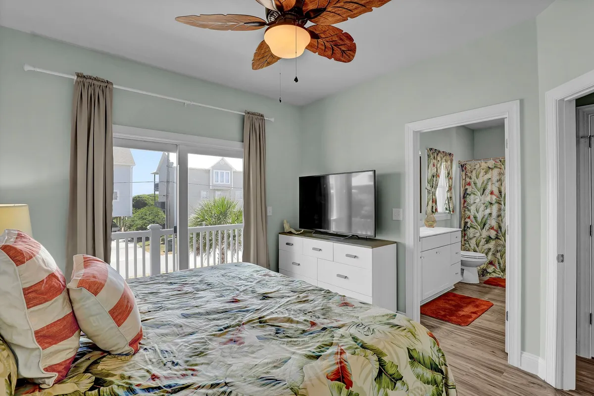 A serene bedroom featuring a bed with tropical bedding, a ceiling fan with a leaf design, a large window letting in abundant natural light, and a television unit. The overall atmosphere is calm and inviting, ideal for unwinding after a day out.