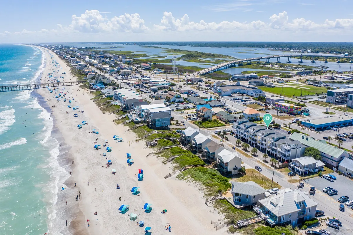 A vibrant scene featuring the beach with colorful umbrellas, a lively pier stretching into the ocean, and town buildings in the background. The area is bustling with people swimming, sunbathing, fishing from the pier, shopping, and enjoying local restaurants. The overall mood is cheerful and social, highlighting both the beach and community attractions.