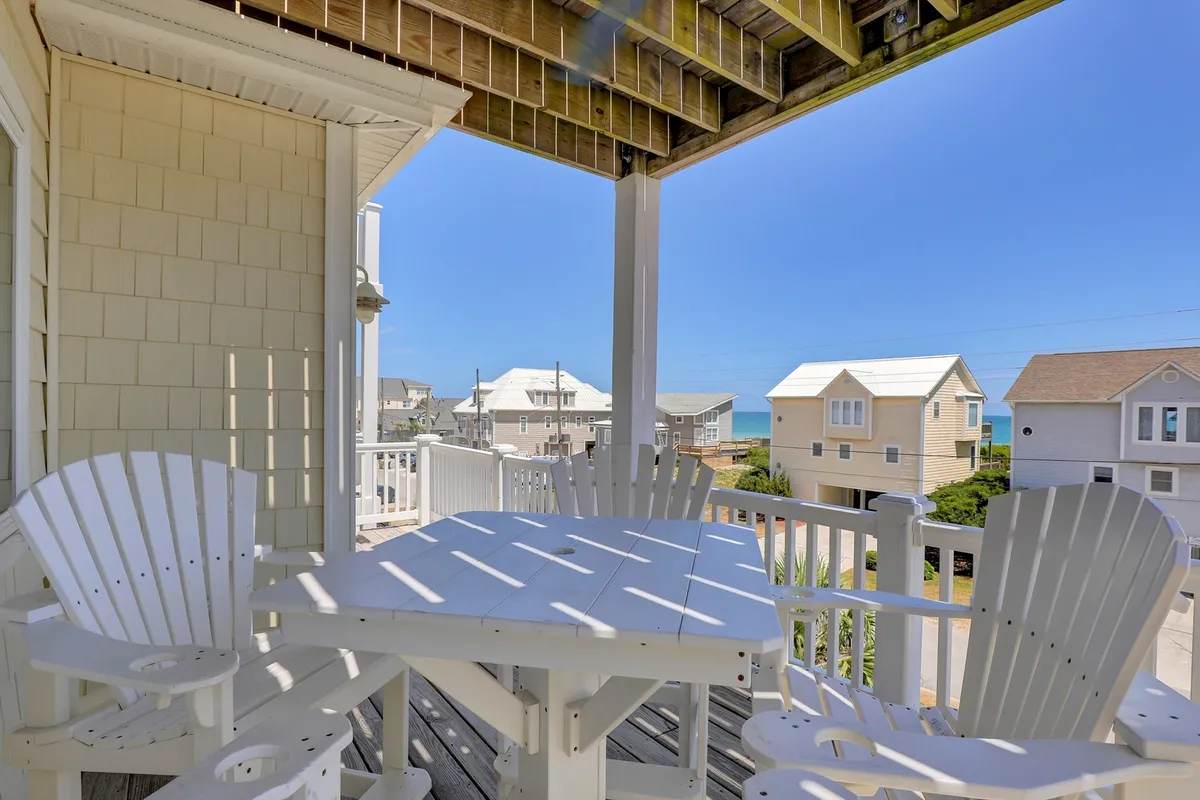 A bright outdoor dining area featuring an inviting table surrounded by adirondack chairs, set against a backdrop of distant beach houses and a clear blue sky. The space feels perfect for group gatherings, al fresco meals, and relaxed conversations as the sun sets. 