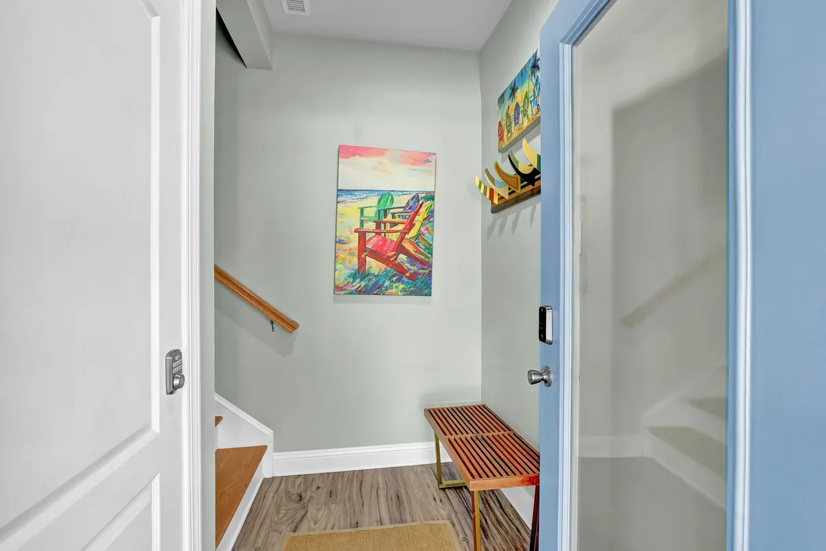 An inviting entryway featuring colorful artwork, a wooden bench for seating or storing beach gear, and a staircase leading into the home. The natural light and cheerful decor create a welcoming first impression for arriving guests.