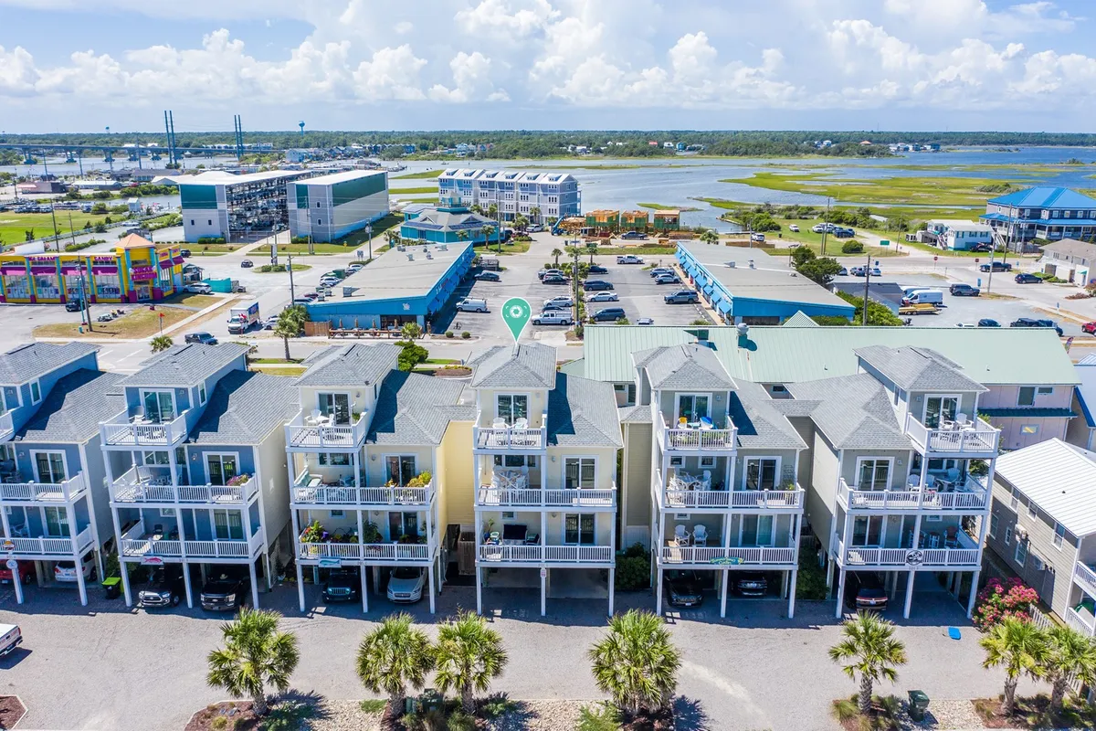 A cheerful exterior shot of multi-story beach houses and palm trees with a parking area and distant water. The scene is filled with light, suggesting a welcoming community vibe. Families gather outdoors, and colorful buildings signal nearby attractions and activities.