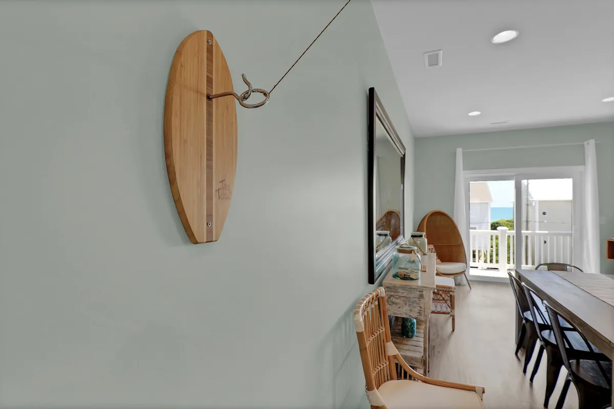 A casual yet stylish dining area featuring a large wooden table, ample seating, and contemporary surfboard wall art. Sunlight pours in through wide windows, highlighting the relaxed, coastal ambiance and inviting communal dining experiences.